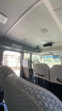 interior view of a passenger van or minibus showing rows of gray patterned seats, overhead AC vents, ceiling grab handles, and a dashboard-mounted camera or sensor device
