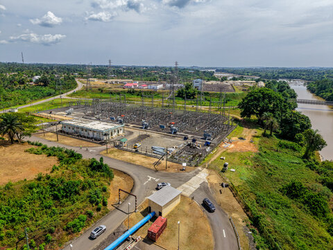 Aerial view of a power substation with electrical transformers and switchyard infrastructure, situated near a river with surrounding green vegetation and roads in Monrovia, Liberia.