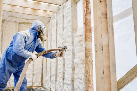 Worker spraying polyurethane foam insulation onto interior wall of timber frame house. Protective respirator, renovation work, thermal sealing and energy efficient building concept