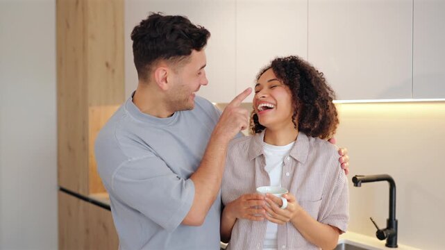 Diverse couple young women and men with coffee in the morning share playful moment in modern kitchen. Man touches her nose affectionately while she holds cup and laughs joyfully.