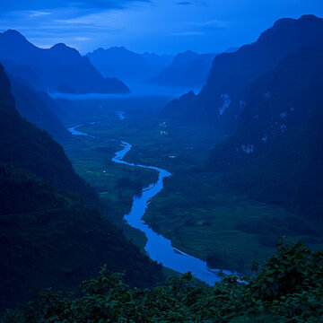 Winding blue river cuts through a misty, mountainous valley at twilight, flanked by terraced green fields and distant peaks