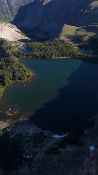 Vertical Screen: Beartooth Highway, Montana, USA drone footage reveals stunning alpine lake surrounded by rugged mountain peaks and evergreen forests in pristine wilderness landscape.