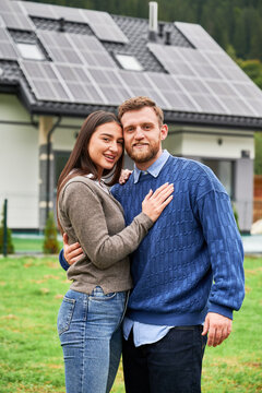 Couple embracing near house with solar panels, smiling at camera. Happy homeowners using renewable energy, eco friendly home and sustainable lifestyle concept