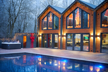 Fototapeta premium Luxury wooden cabins glow beside outdoor pool at winter dusk. Festive lights, panoramic windows and warm interiors create cozy holiday resort atmosphere