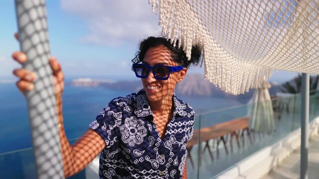 Smiling woman wearing blue sunglasses relaxes under woven net canopy on glass terrace overlooking caldera sea views in Santorini Greece. Warm golden sunlight creates pattern shadows on her face.