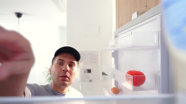 Young man in black cap peers into nearly empty refrigerator searching for food, finding only a single red tomato and egg on door shelves in minimalist modern white kitchen with pendant lighting.