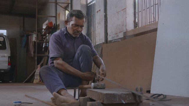 An indoor close-up shot of a skilled Indian Asian male worker or craftsman sitting, checking alignment and striking the hammer with precision to straighten and correct the shape of the metal rod.