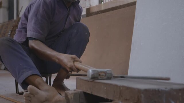 An indoor close-up shot of a skilled Indian Asian male worker sitting, checking alignment and striking the hammer with precision to straighten the shape of the metal rod in a fabrication workshop