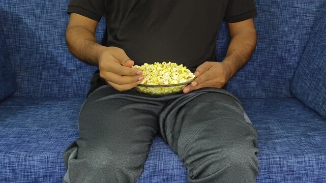 Adult lazy depressed person in casual attire sitting on sofa holding bowl of popcorn in hand for eating while watching weekend movie streaming on OTT. Online digital entertainment industry concept.