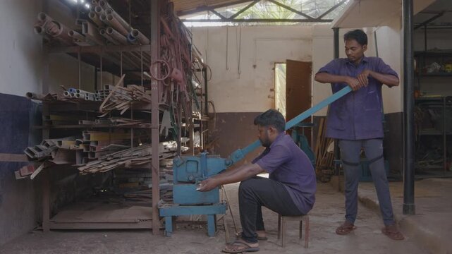 A close indoor shot of two men or male skilled workers or craftsmen, working together, precisely cutting a metal rod into different shapes using a manual cutting machine in a fabrication workshop