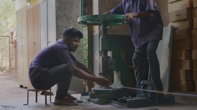 A mid indoor shot of two Indian Asian male skilled workers or craftsmen precisely bending a metal rod into shape using a manual bending machine in a fabrication workshop. 