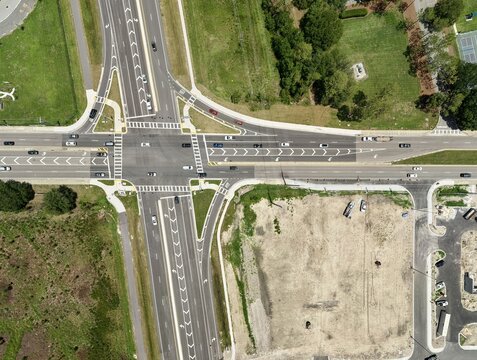 Aerial view of a road intersection with vehicles, surrounding green spaces, and a cleared construction site in the United States.