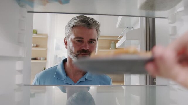 Close-up footage of gray-haired man with beard opening empty refrigerator. Hand from fridge putting sandwich on shelf. Happy man picking up food and closing door. Modern kitchen in background.
