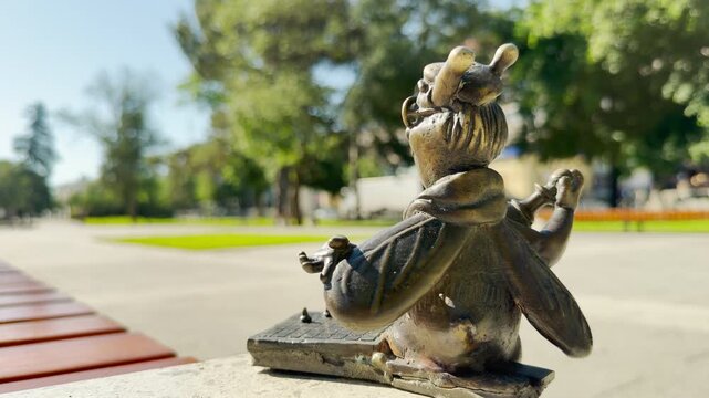 A charming bronze bee figurine overlooks a sunny park from a stone ledge in Simferopol, Crimea. The playful miniature scene stirs a warm feeling of calm delight. Green trees fill the background.