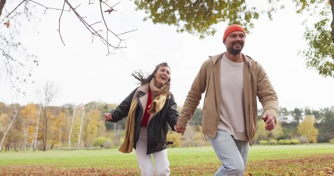 Couple running in autumn park, smiling outdoors. Warm jackets, scarves and hats show seasonal style as golden leaves create a bright warm backdrop. Concept of love, happiness and togetherness.
