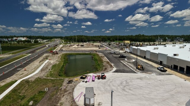Aerial view of a construction site featuring a large warehouse building, a retention pond, heavy machinery, and a paved road under a blue sky in the United States.
