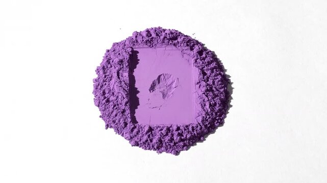 Vertical crane view of a heap of violet powder with a rectangular sign on a dim grey background, lit with orbiting light causing moving shadows