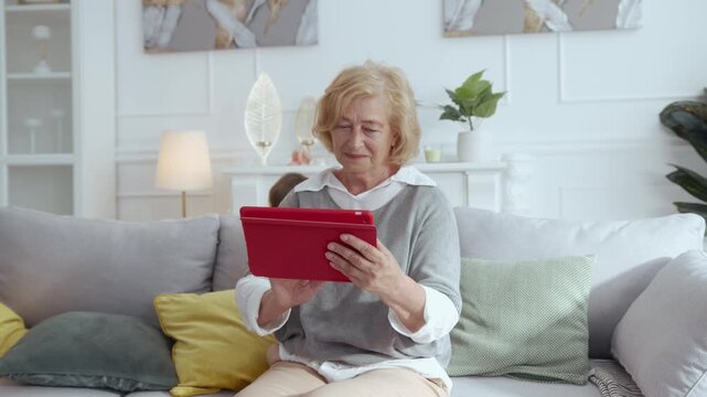 An elderly woman is comfortably sitting on a cozy couch using a tablet computer