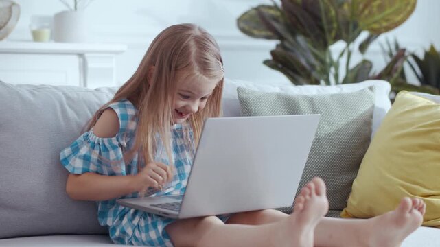 A little girl is comfortably sitting on a couch using her laptop computer