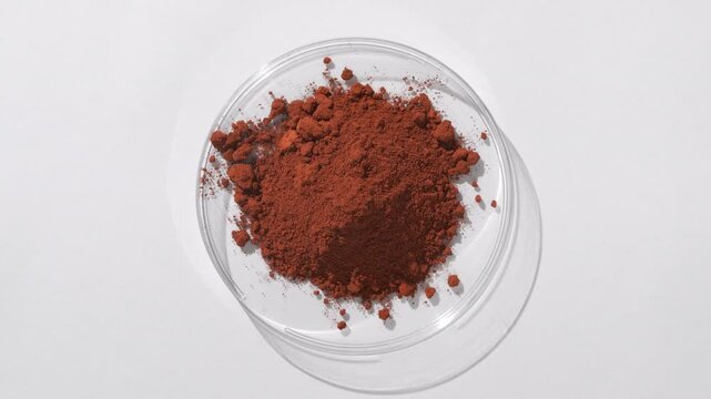 Top view of a mountain of brown fine powder in a plastic petri dish on a grey surface, illuminated by rotating light casting evolving shadows | Black tea powder cosmetic ingredient concept