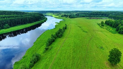Obraz premium Lush green fields adjacent to a calm river reflecting cloudy skies and trees in the distance