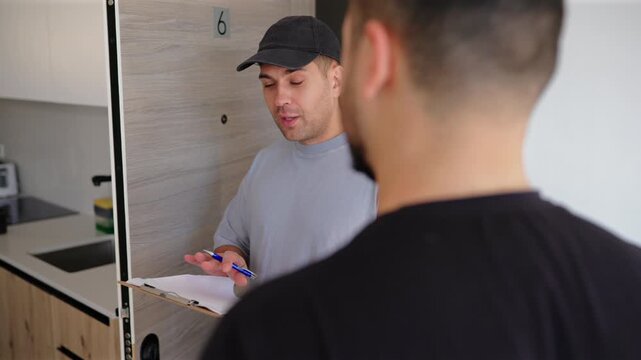 Delivery worker wearing gray t-shirt and black cap holds clipboard while speaking with customer at modern apartment doorway number six, taking notes during friendly doorstep conversation.