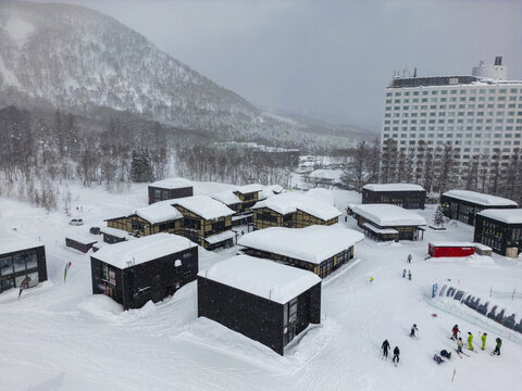 Aerial view of Niseko ski resort with modern dark-timber buildings covered in deep snow, surrounded by snow-covered trees and mountains, with skiers on the slopes in Niseko, Hokkaido, Japan.