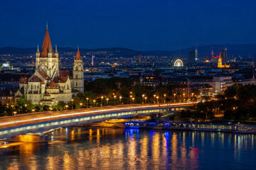 Fototapeta premium Beautiful evening city skyline with illuminated historic church bridge river reflections and urban lights at blue hour 