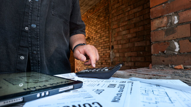 Construction professional calculating costs on a calculator with blueprints and tablet