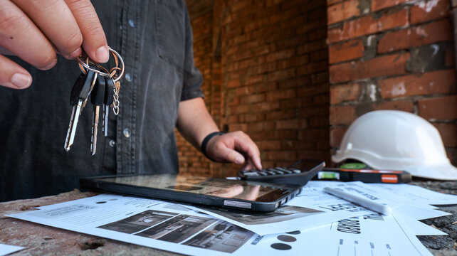 Construction worker holding keys and using calculator with blueprints and hard hat nearby