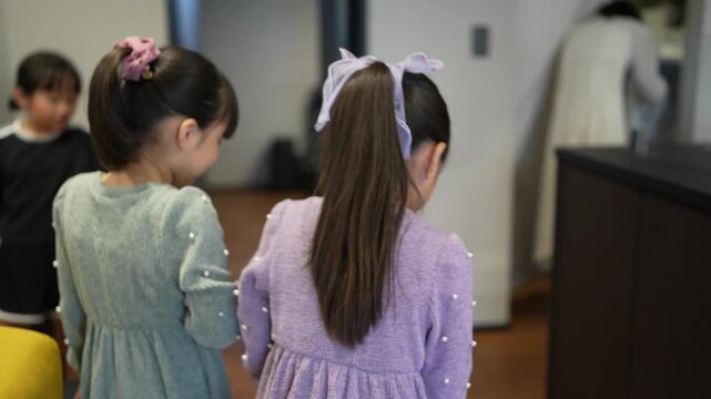 Two young girls stand side by side, watching something ahead. One wears a lavender dress, the other a gray sweater. Both have hair adorned with bows or clips. Their attention is fixed forward, curious