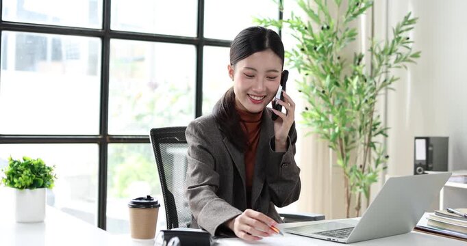 Asian businesswoman working in a professional office, multitasking with a laptop and smartphone while reviewing documents, embodying efficiency and modern corporate communication
