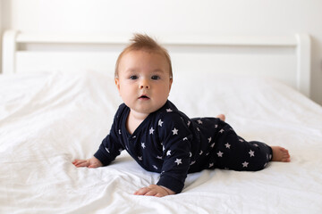 Fototapeta premium A cute little caucasian six-month-old baby boy, dressed in a black jumpsuit with stars, is crawling on all fours on a bed with white linens. Lifestyle close-up portrait in home setting. Real child. 
