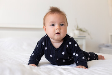 Fototapeta premium A cute little caucasian six-month-old baby boy, dressed in a black jumpsuit with stars, is crawling on all fours on a bed with white linens. Lifestyle close-up portrait in home setting. Real child. 