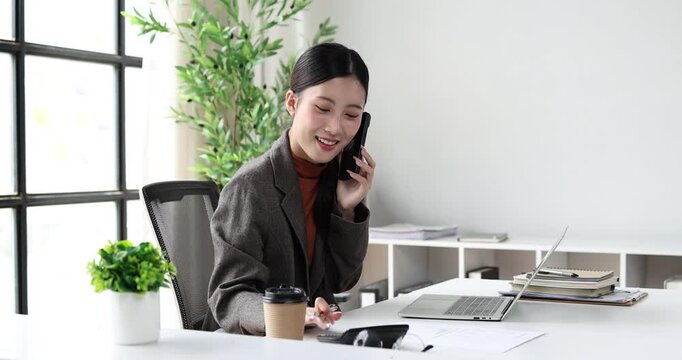 Asian businesswoman working in a professional office, multitasking with a laptop and smartphone while reviewing documents, embodying efficiency and modern corporate communication