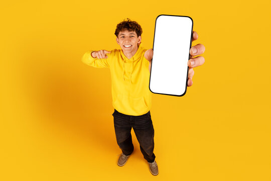Smiling young guy in yellow hoodie holding blank smartphone screen toward camera on yellow studio background for app mockup or mobile promotion