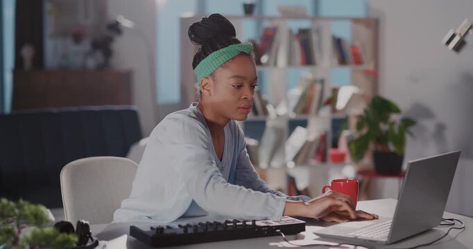 Busy positive african young woman in casual outfit wearing headphones playing electronic piano keyboard recording music using online application technology.