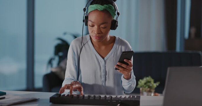 Young woman artist musician recording music tracks with electronic piano keyboard synthesidzer using mobile phone listening in headphones. African ethnicity. Apartment.