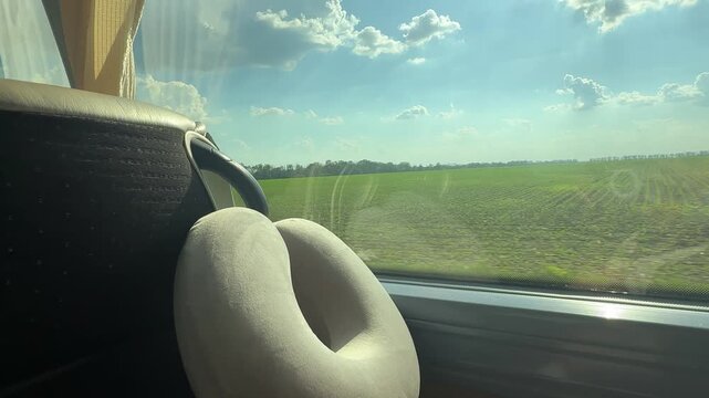 Travel neck pillow resting beside a bus window overlooking a green countryside landscape. Comfortable road travel, public transportation, tourism, relaxation and journey concept.