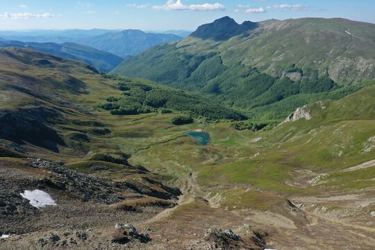 Aerial view of Lago della Bargetana nestled in the green valleys of the Apennine Mountains, surrounded by rocky slopes and patches of snow in Ventasso, Emilia-Romagna, Italy.
