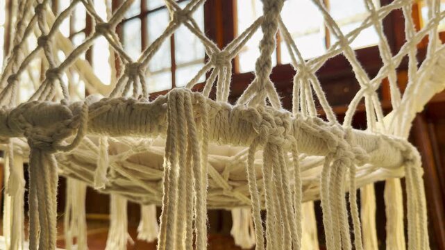 A cozy white macrame hammock chair hangs in a sunlit room. Its intricate woven cords and fringe create a warm, peaceful, and relaxing atmosphere, perfect for a quiet afternoon.