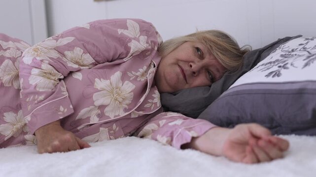 Senior Woman Resting in Bed. Senior woman rests in bed at home, opens her eyes and gently touches her face while lying under a blanket.
