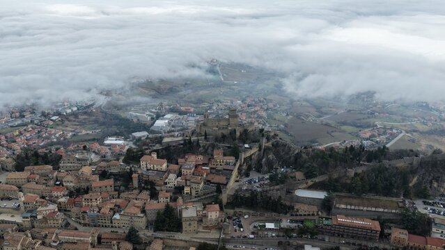 Aerial view of Guaita Tower perched on a rocky ridge above the historic old town, surrounded by a sea of clouds and rolling hills. Citta di San Marino, San Marino, San Marino.