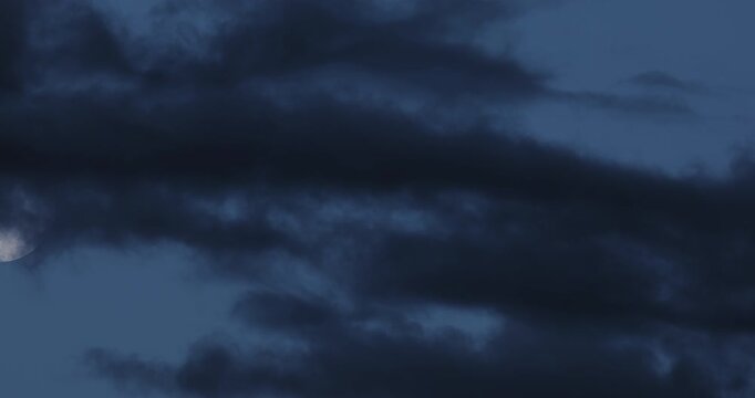 Dark storm clouds race across a deep blue night sky as a three-quarter illuminated moon with sharp visible craters drifts between them. Dramatic cinematic timelapse for films, trailers and commercials