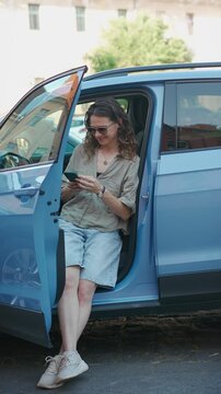 Young female driver using a smartphone while standing next to a parked car on a city street in Europe, representing modern mobility and connected lifestyle.