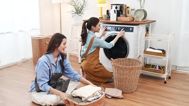 Two women folding laundry together bright laundry room beside front load washing machine and wicker basket, young woman and friend organizing folded clothes home cozy domestic laundry room with