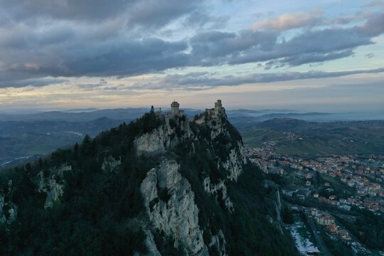 Aerial view of the historic towers of San Marino perched on the rocky cliffs of Mount Titano with surrounding landscape under a cloudy sky in San Marino, San Marino, San Marino.
