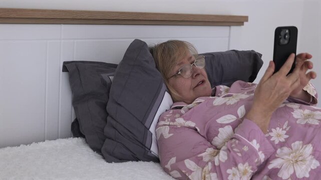 Senior Woman Scrolling on Smartphone in Bed. Senior woman lying in bed at home and scrolling on a smartphone. Mature woman in pajamas browses content on her mobile phone while resting.