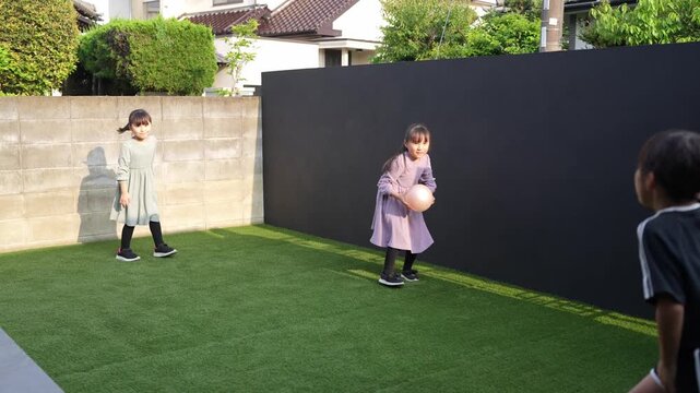 Three young girls play on artificial grass. One girl chases a pink ball. Another kneels near a tall black wall. Third girl runs toward the ball. They laugh and play together