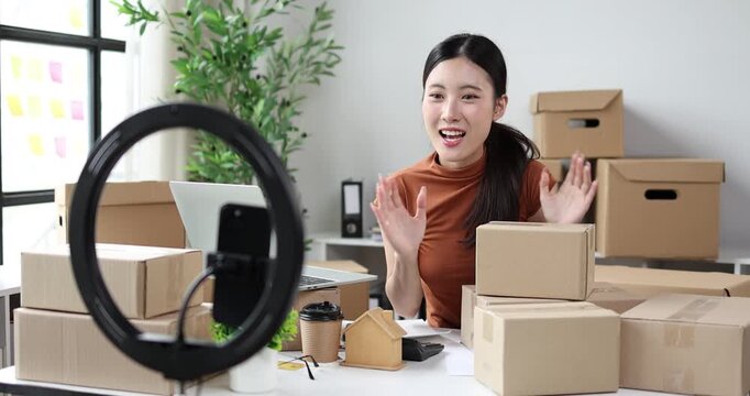 Young asian woman livestreaming e-commerce sales using a smartphone and ring light, receiving customer orders and preparing shipping packages from her home office small business